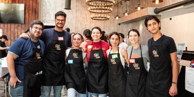 Group of volunteers wearing matching aprons and name tags, standing together inside a community event space with tables and workstations in the background.