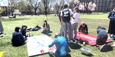 People gathered on a grassy lawn creating large painted artworks on canvases spread across the ground, with art supplies, trees, and campus buildings in the background.