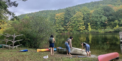 Three people standing beside canoes on the shore of a calm lake, surrounded by forested hills and trees reflected in the water.
